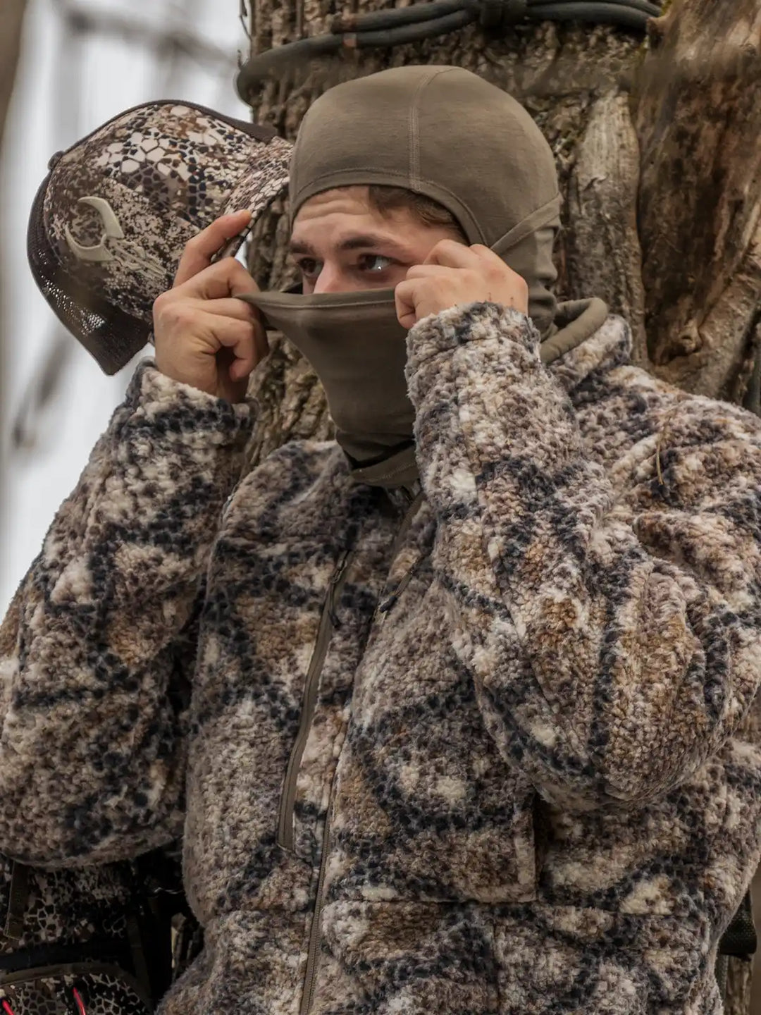 male hunter pulling neck gaiter over face, sitting on tree stand