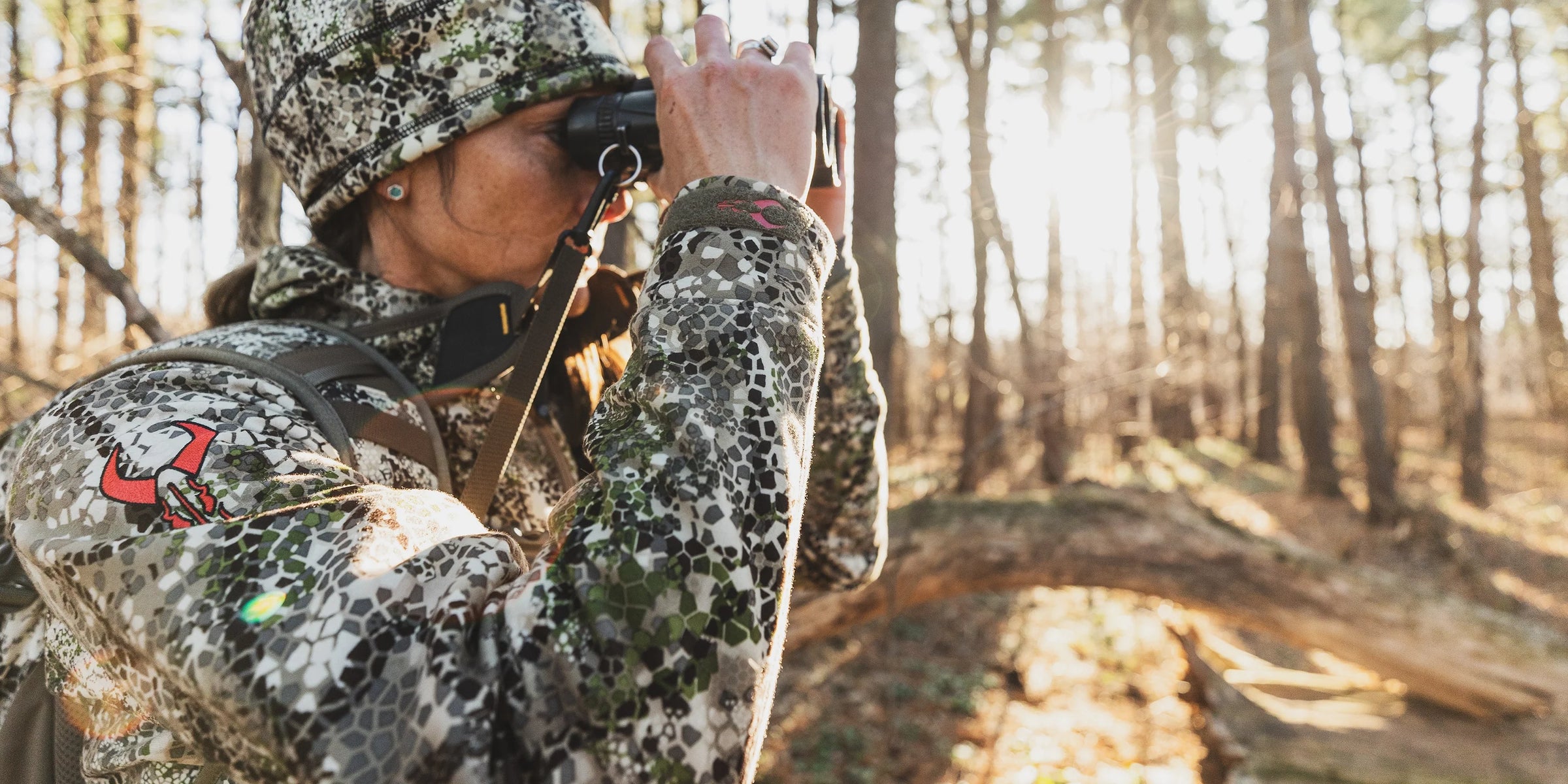 Person in Badlands camo gear with skull logo uses binoculars in a sunlit forest, highlighting outdoor activity and brand.