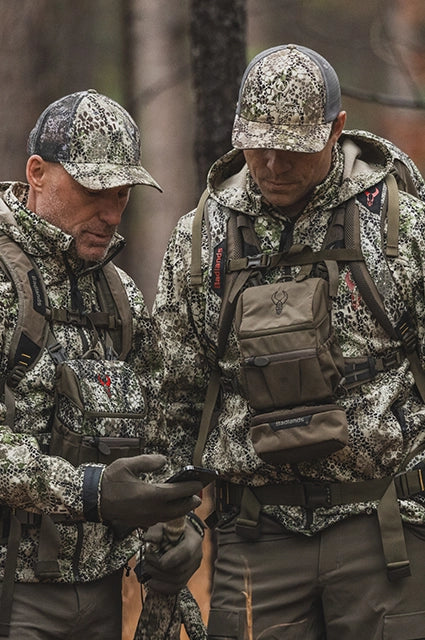 Two hunters in camo gear wearing Badlands backpacks with skull logo, standing outdoors and looking at a handheld device.