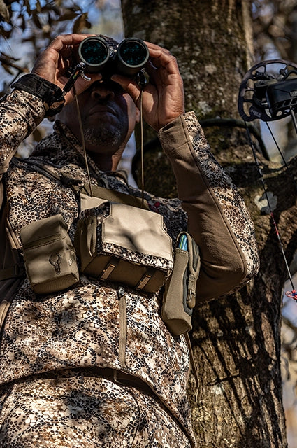 Hunter in Badlands camo uses binoculars; Badlands logo skull visible on chest pouch. Bow hanging on tree in background.