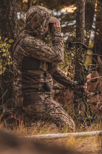 Hunter in Badlands camo kneels in forest, holding a bow and using binoculars. Badlands skull logo visible on gear.