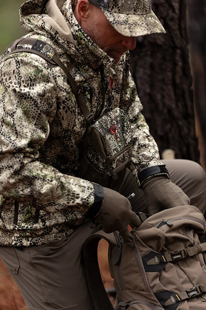Hunter in camo gear with Badlands logo on chest pouch, adjusting a tan backpack outdoors.