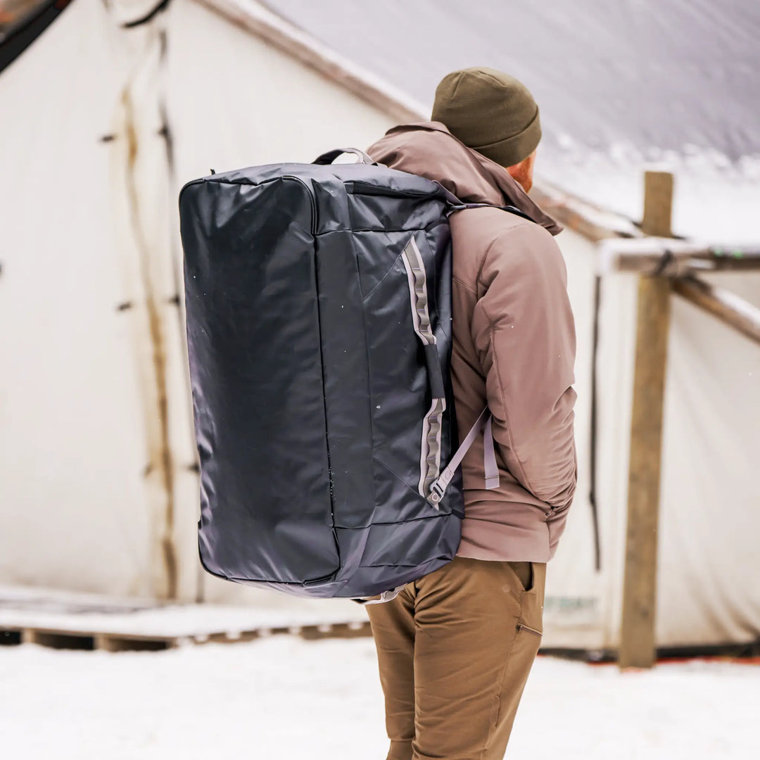 Person wearing a large Badlands duffel backpack outdoors in snowy conditions; tent and gear visible in background.
