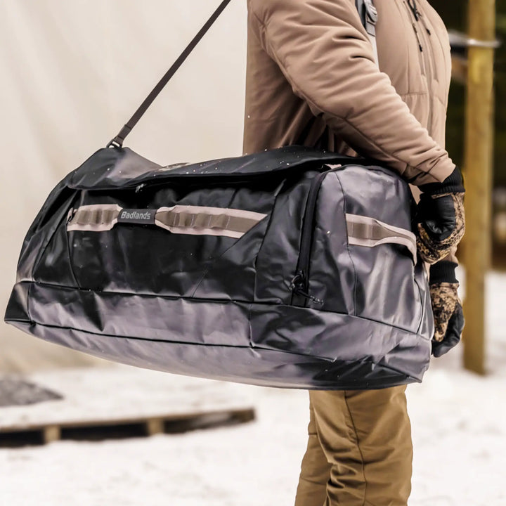 Person carrying a large Badlands duffel bag outdoors; Badlands skull logo visible on the bag.