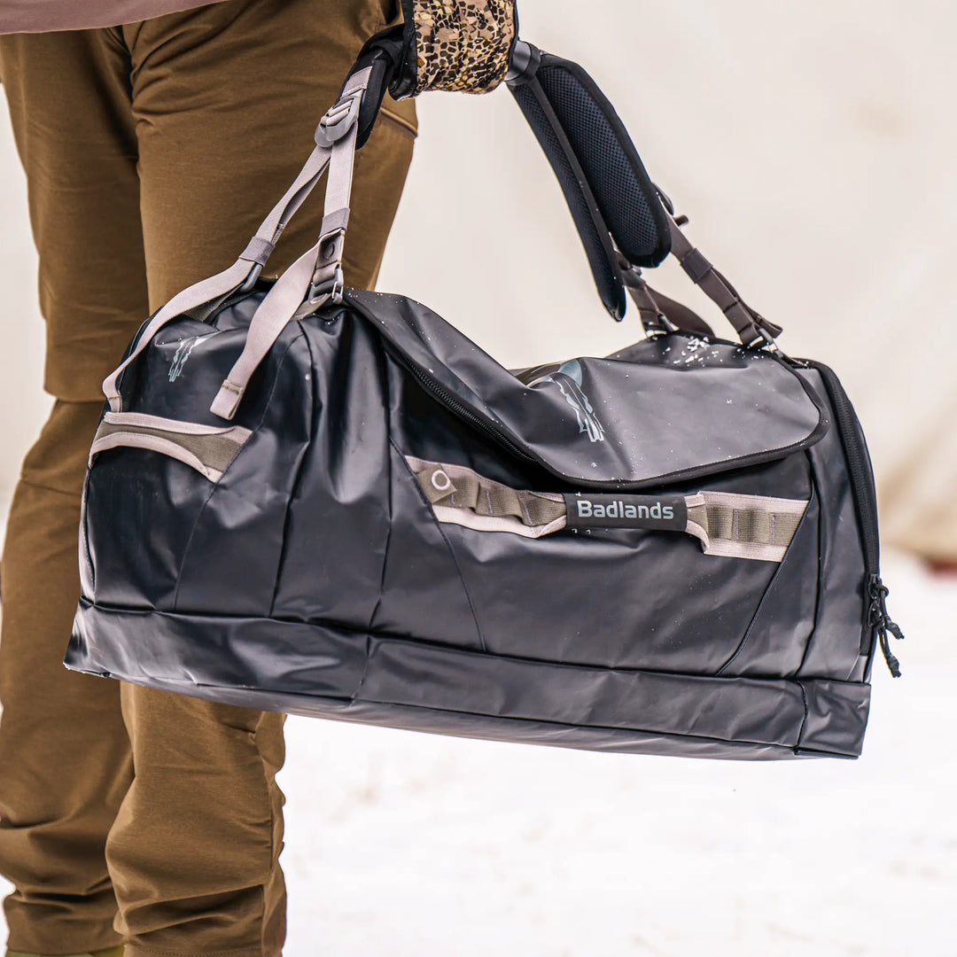Person holding a black Badlands duffel bag with logo skull, outdoors on snow. Bag features sturdy handles and side straps.