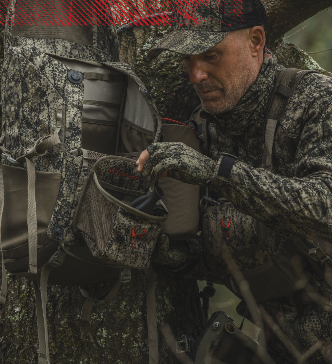 Person in Badlands camo gear accesses a camo backpack with Badlands skull logo, outdoors near a tree.