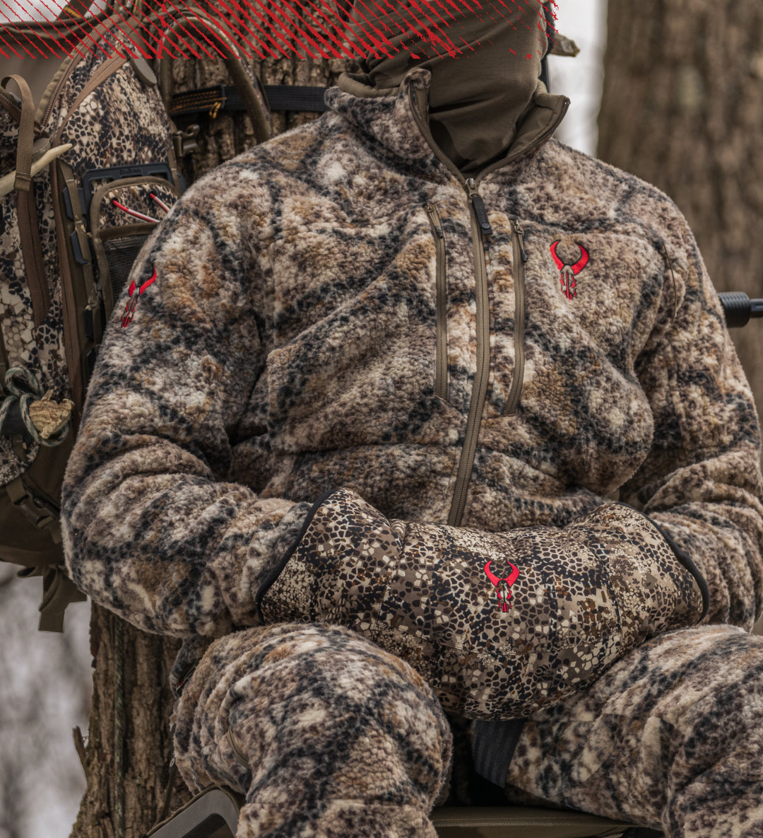 Hunter in Badlands camo fleece with red Badlands skull logo, sitting outdoors with matching mitt and gear in a tree stand.