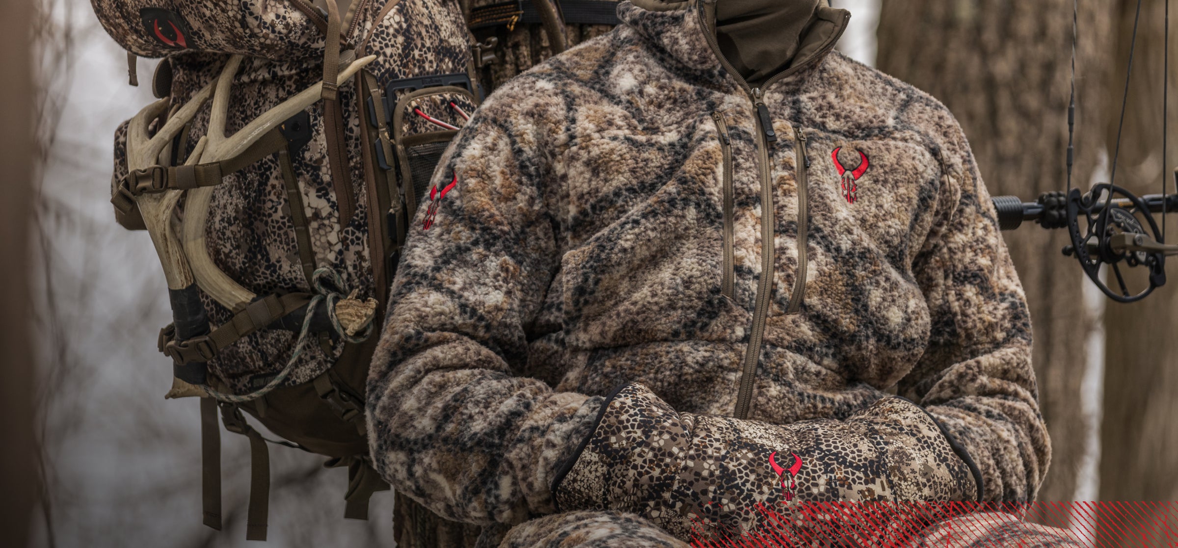 Hunter in Badlands camo gear with red skull logo, holding bow; antlers strapped to matching backpack in a forest setting.