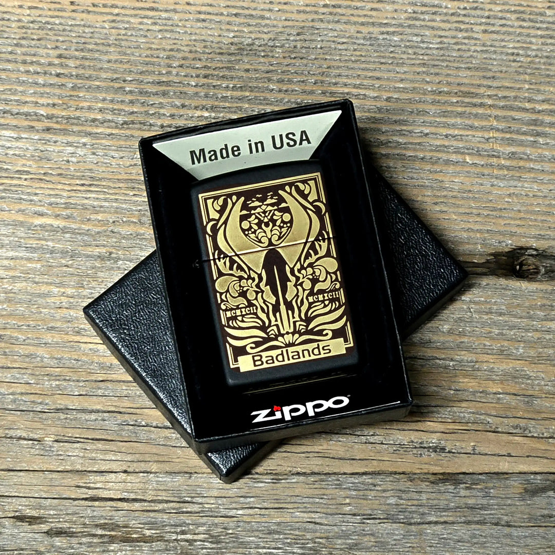 Gold Zippo lighter with Badlands skull logo and antlers, in a black box labeled "Made in USA," on a wood surface.