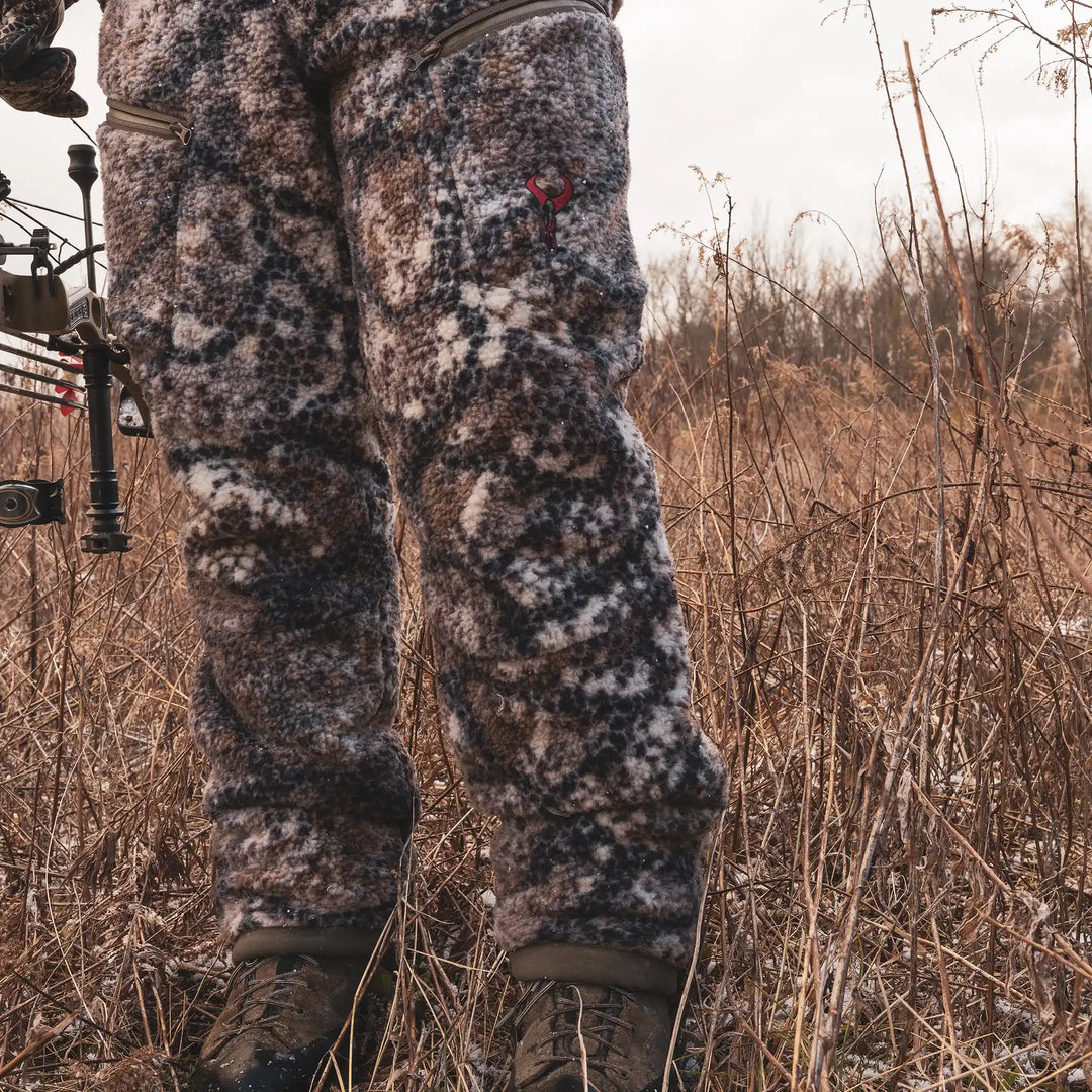 Hunter in Badlands camo fleece pants with red skull logo, standing in dry grass, holding a bow.