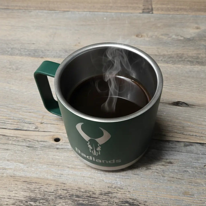 Green Badlands mug with skull logo, filled with steaming coffee, sits on a wooden surface.