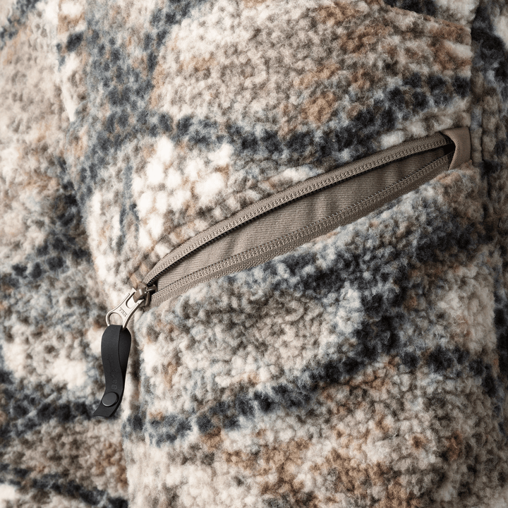 Close-up of Badlands fleece jacket pocket with zipper and skull logo pull, in earth-tone camo pattern.