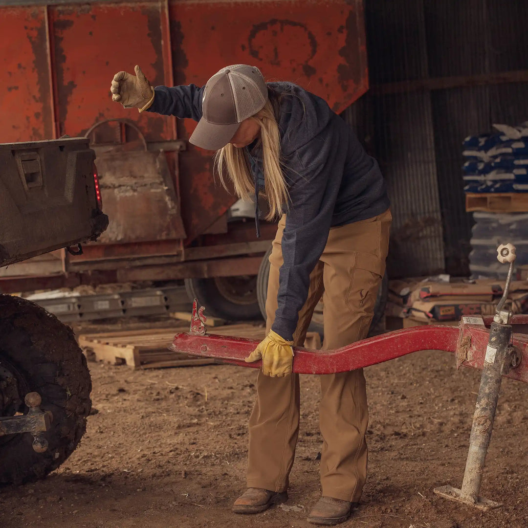 Woman in Badlands pants and gloves hitches a trailer in a barn; Badlands skull logo visible on her pocket.