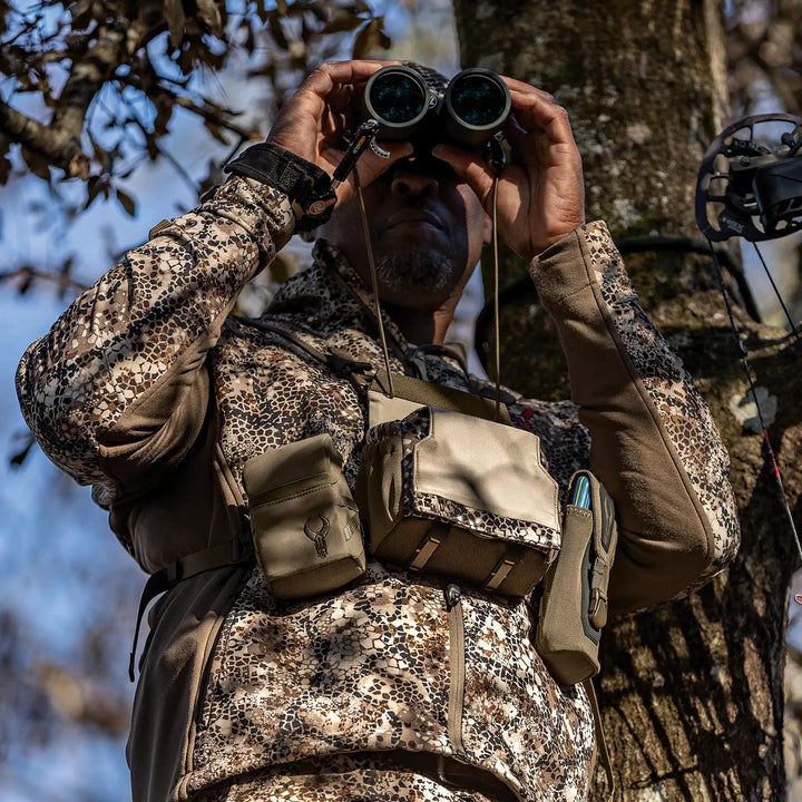 Hunter in Badlands camo uses binoculars; Badlands logo skull visible on gear pouch; tree and bow in background.