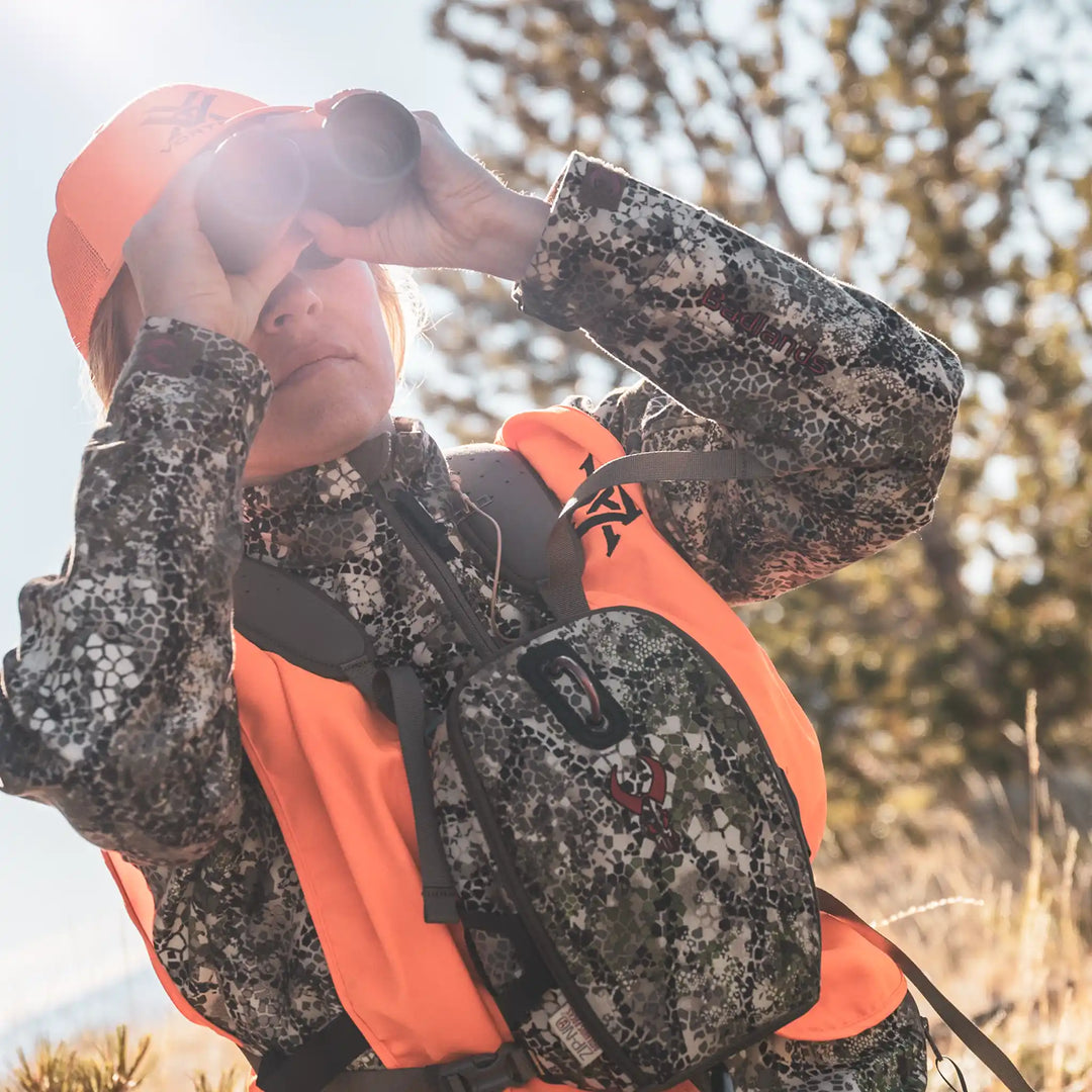 Hunter in camo and orange vest uses binoculars; Badlands logo skull visible on chest pack. Outdoor daylight setting.