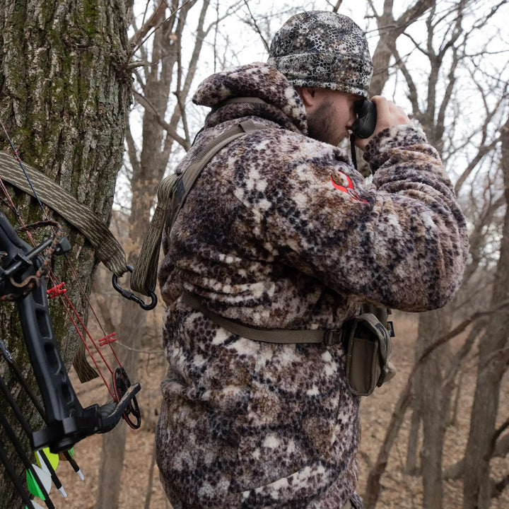 Hunter in Badlands camo gear with skull logo, using binoculars in a tree stand, bow and arrows visible, forest background.