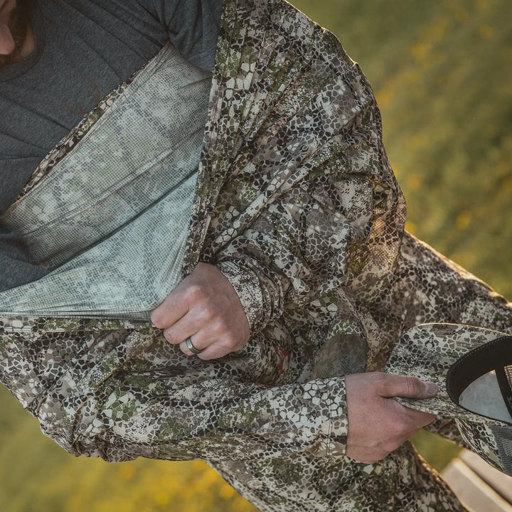 Person wearing Badlands camo jacket, holding matching cap with Badlands skull logo, outdoors in natural light.