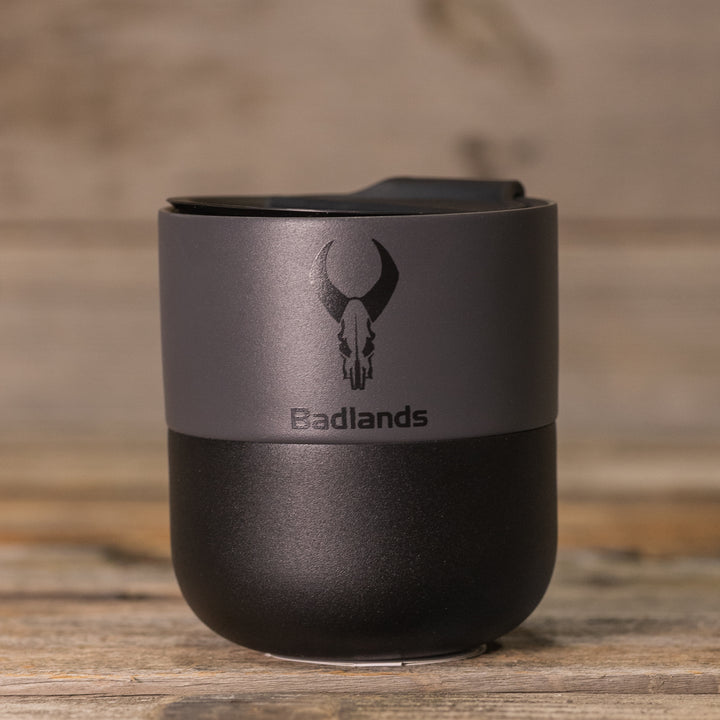 Black and gray Badlands travel mug with skull logo, placed on a wooden surface.