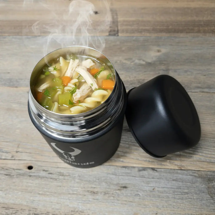 Hot chicken noodle soup in a Badlands thermos with logo skull, lid off, on a wooden table.