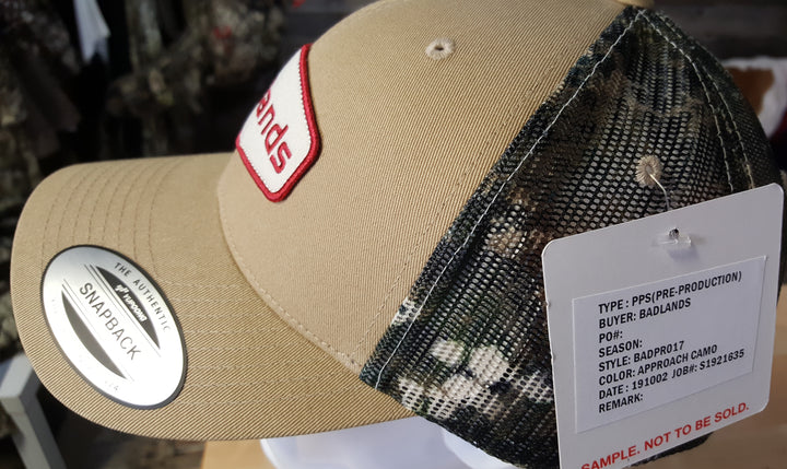 Tan and camo Badlands snapback hat with mesh back, logo patch, and sample tag; not for sale.