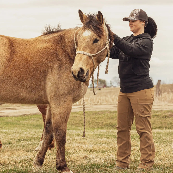 Person in Badlands hoodie and logo cap stands beside a brown horse in a grassy field, adjusting the horse's rope halter.
