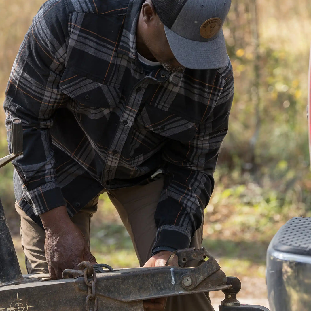 Man in plaid shirt and Badlands logo hat secures trailer hitch outdoors; autumn background.