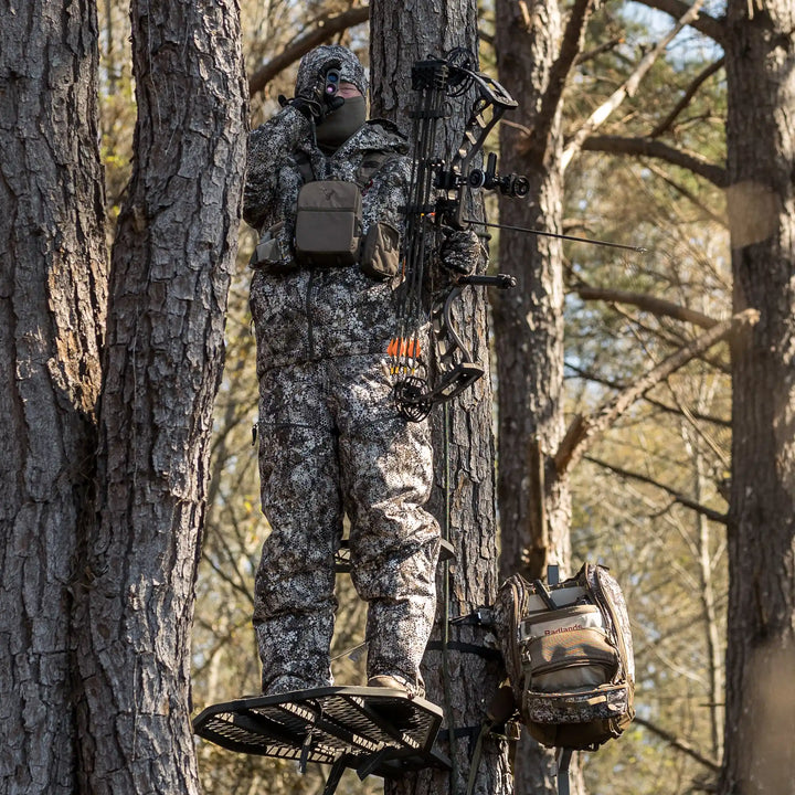 Hunter in camo gear stands on a tree stand with bow, Badlands pack, and logo skull visible; forest background.