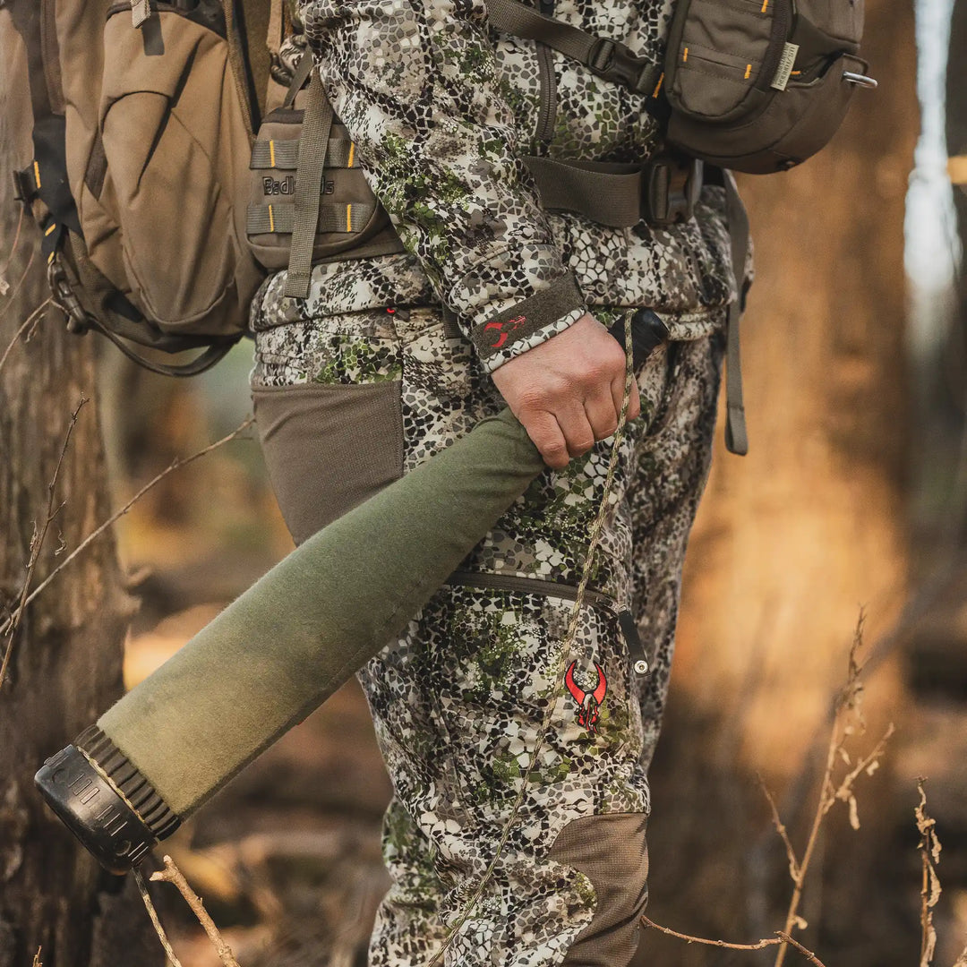 Person in Badlands camo gear holding hunting equipment; Badlands skull logo visible on pants in a forest setting.