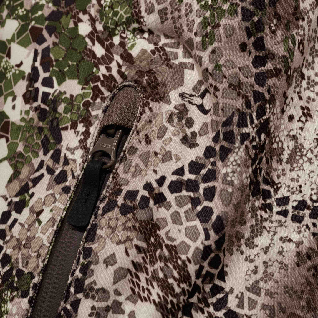 Close-up of Badlands camo fabric with zipper and subtle skull logo, showing textured hexagonal pattern in earth tones.