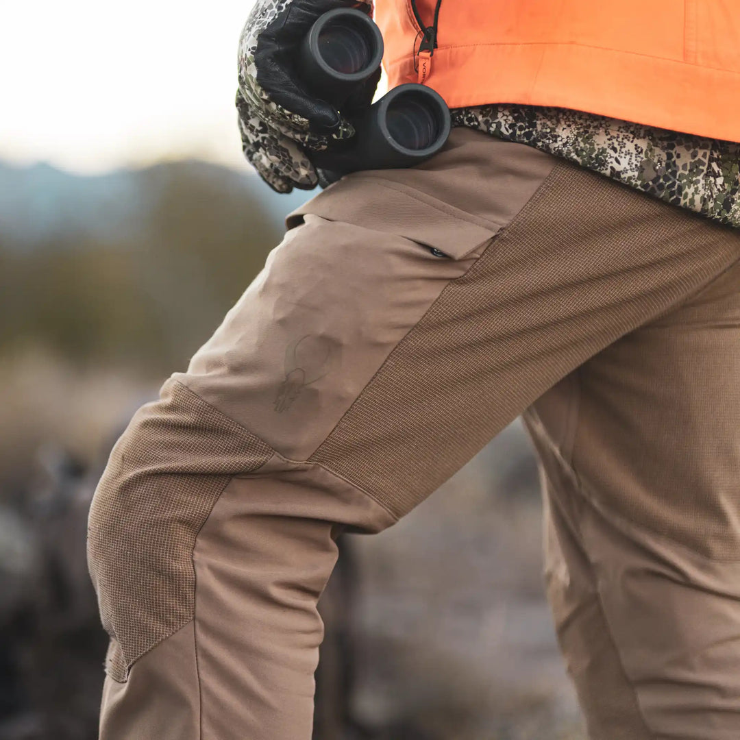 Close-up of Badlands hunting pants with skull logo, person holding binoculars outdoors.