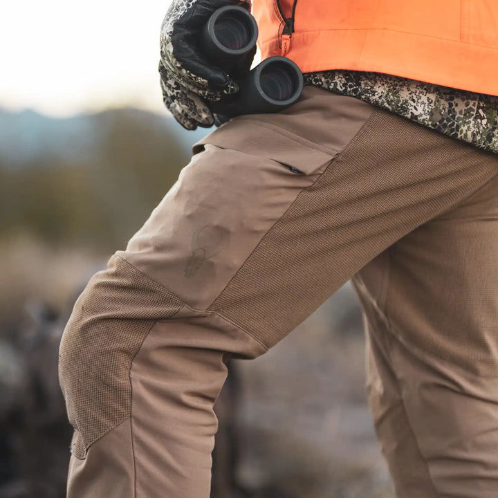 Close-up of Badlands hunting pants with skull logo, person holding binoculars outdoors.