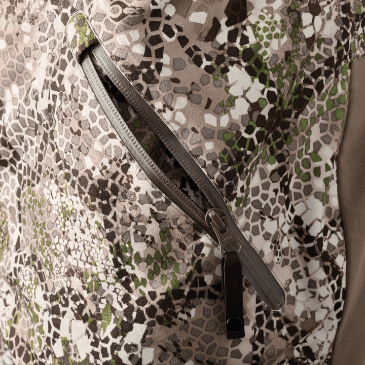 Close-up of a Badlands camo jacket pocket with zipper and subtle Badlands skull logo on the fabric.