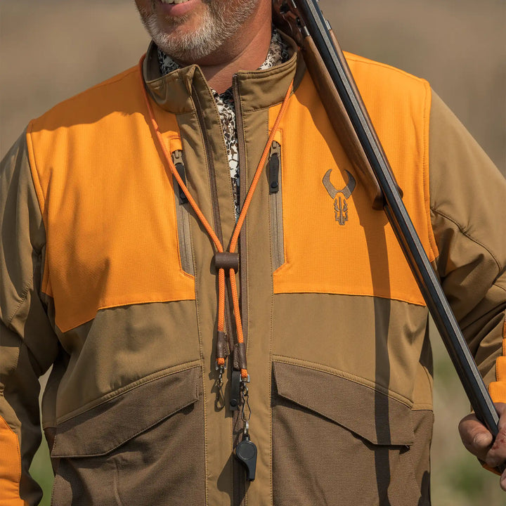 Man in orange and brown Badlands jacket with skull logo, holding a shotgun over shoulder; outdoors, chest view.