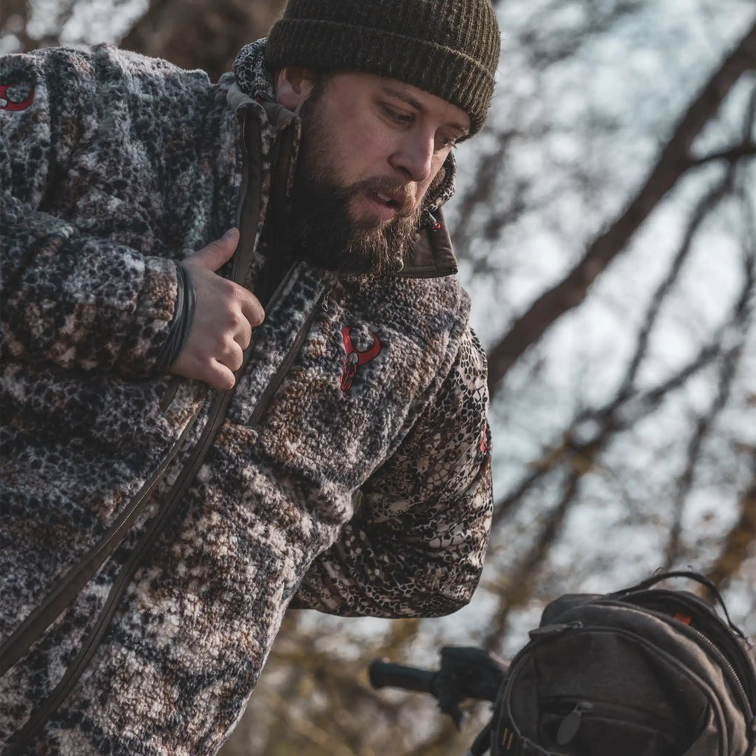 Person wearing a camo Badlands jacket with visible skull logo, outdoors near a backpack and trees in the background.