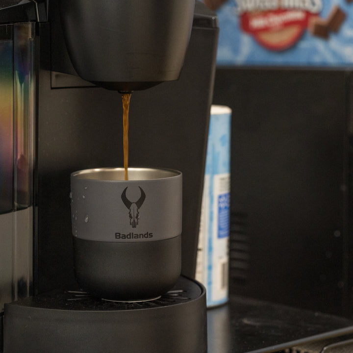 Coffee brewing into a Badlands mug with skull logo, placed under a coffee machine in a kitchen setting.