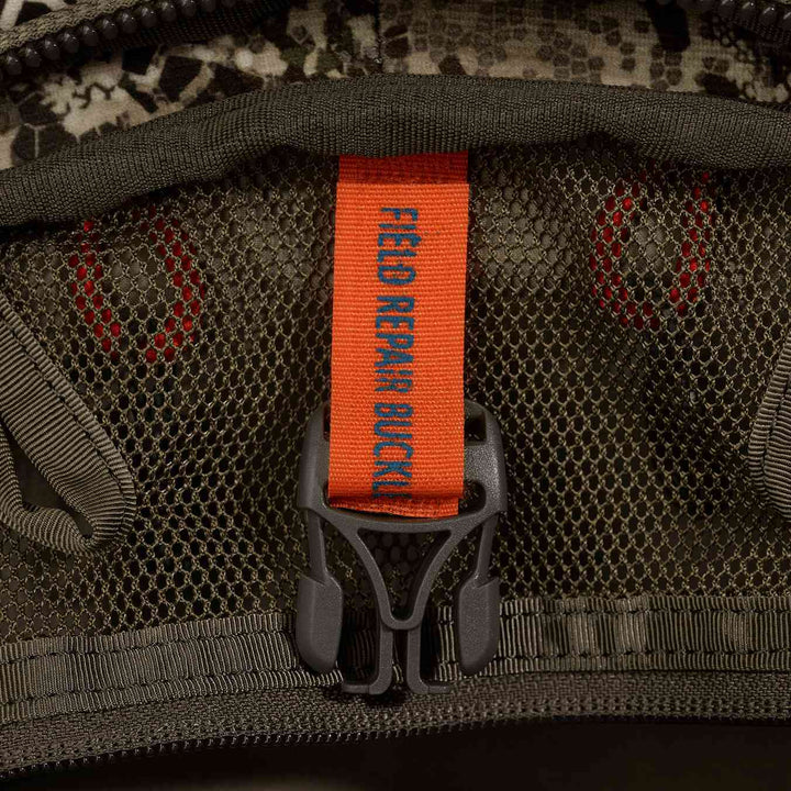 Close-up of a Badlands pack mesh pocket with orange "Field Repair Buckle" tag and Badlands skull logo in background.