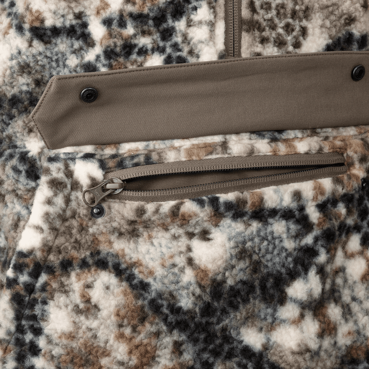 Close-up of Badlands camo fleece fabric with zippered pocket and snap flap; skull logo zipper pull visible.