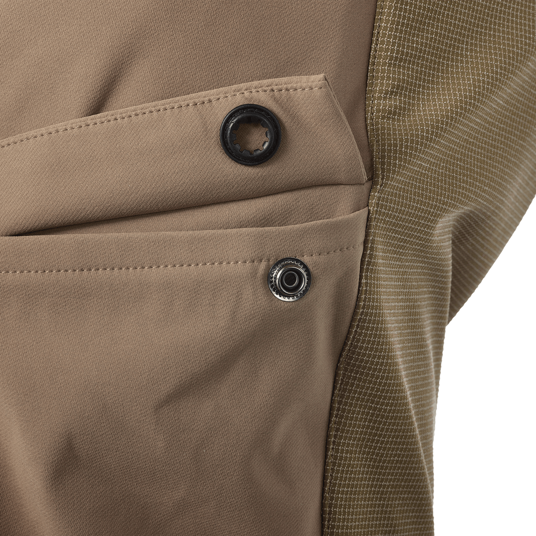 Close-up of tan Badlands pants pocket with logo skull rivet and snap button, showing durable fabric and stitching detail.
