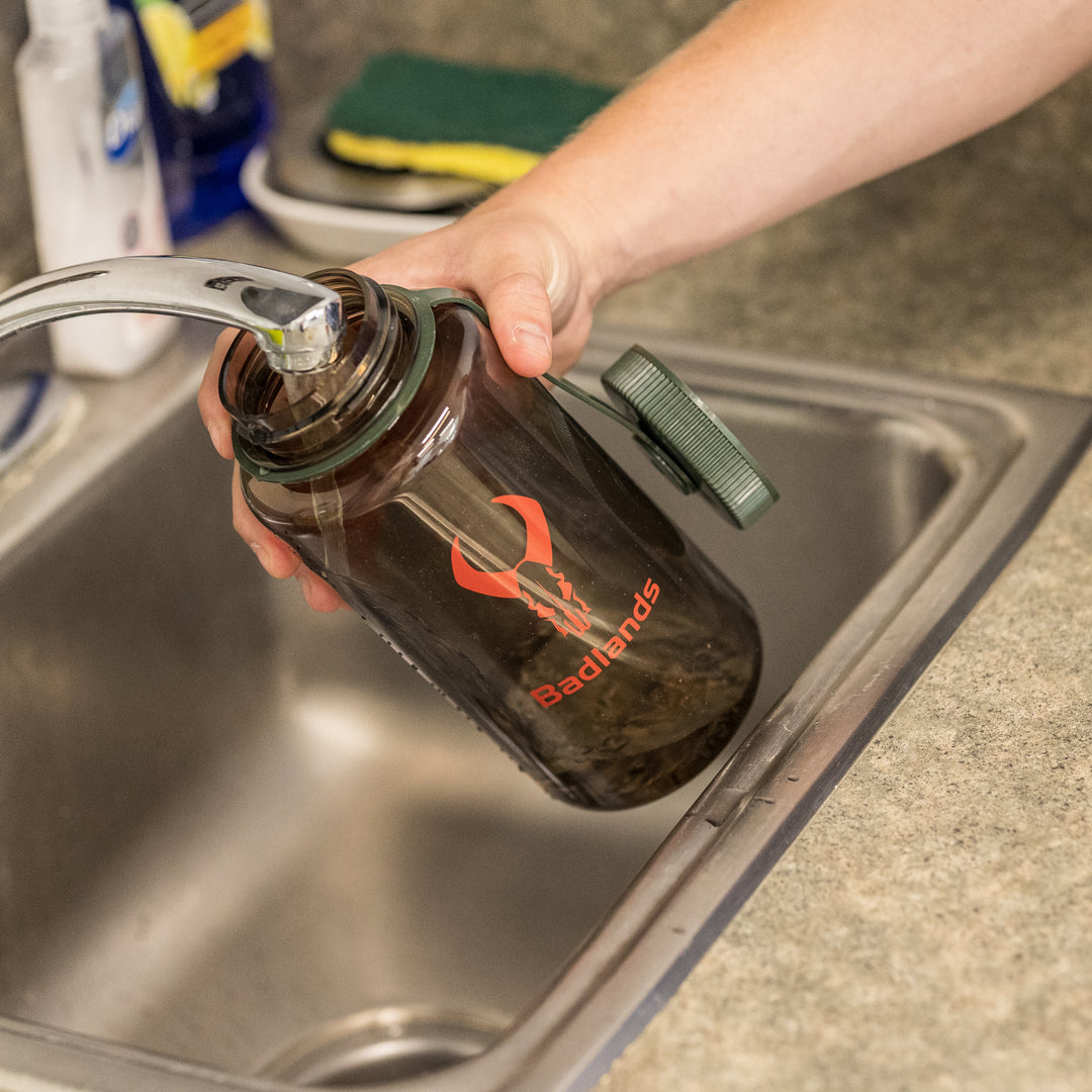 Hand filling a Badlands water bottle with skull logo at a kitchen sink.