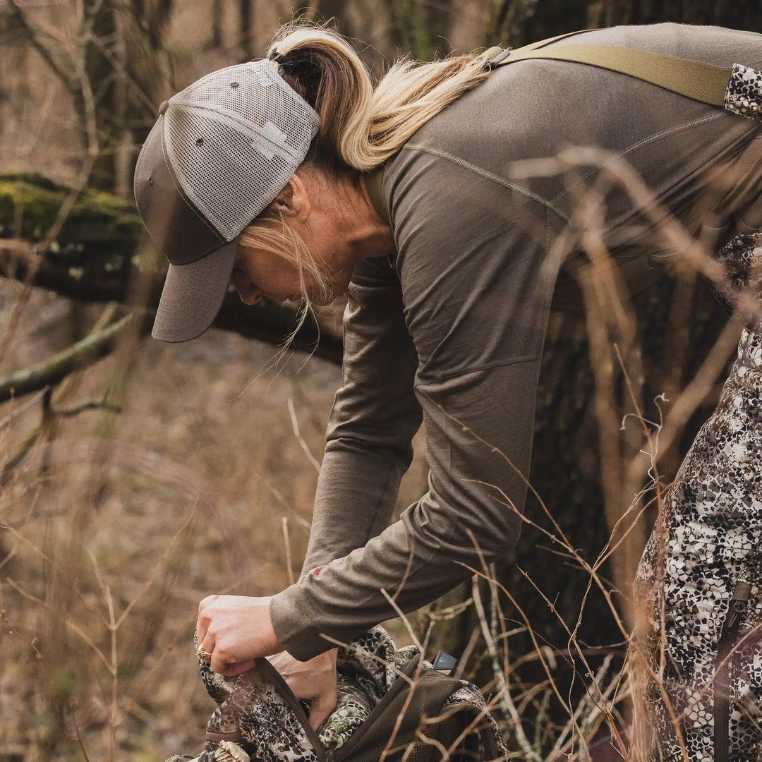 Woman in Badlands camo gear and cap with skull logo, reaching into a camo backpack in a wooded, brush-filled area.
