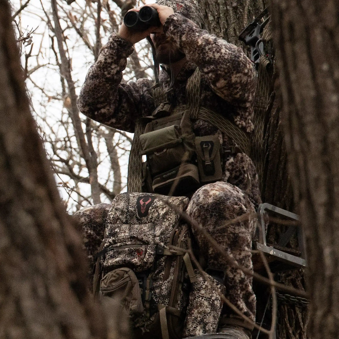 Hunter in camo gear sits in a tree stand, using binoculars. Badlands logo skull visible on camo backpack.