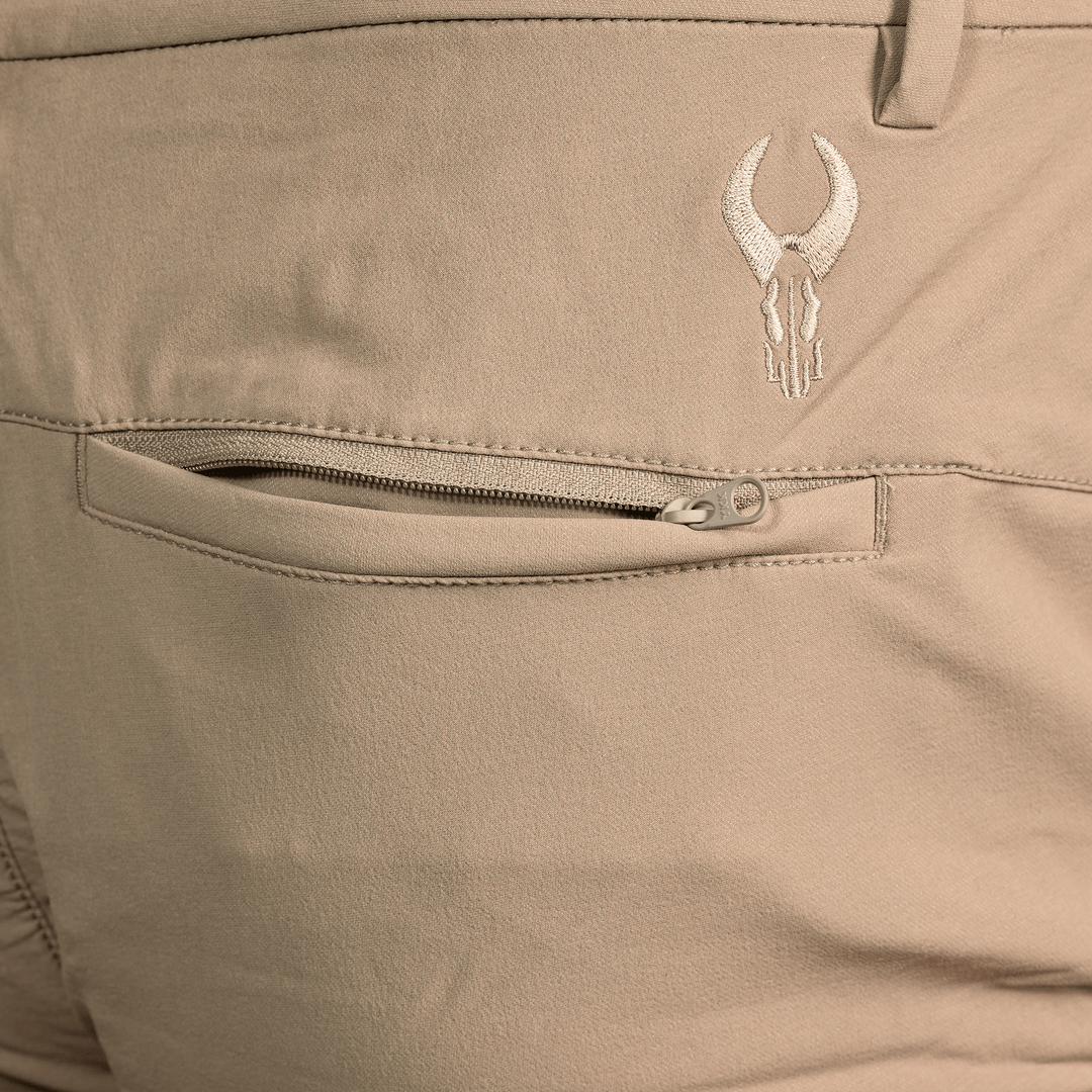 Close-up of tan Badlands pants showing a zippered back pocket and embroidered Badlands skull logo above the pocket.