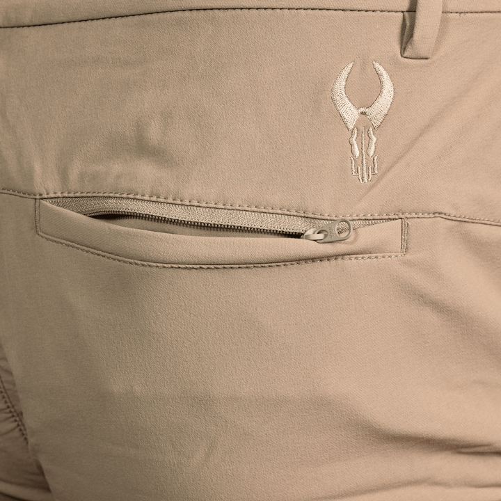 Close-up of tan Badlands pants showing a zippered back pocket and embroidered Badlands skull logo above the pocket.