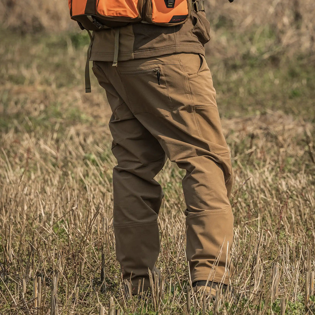 Person wearing tan Badlands pants and an orange backpack stands in a grassy field. Badlands skull logo visible on gear.