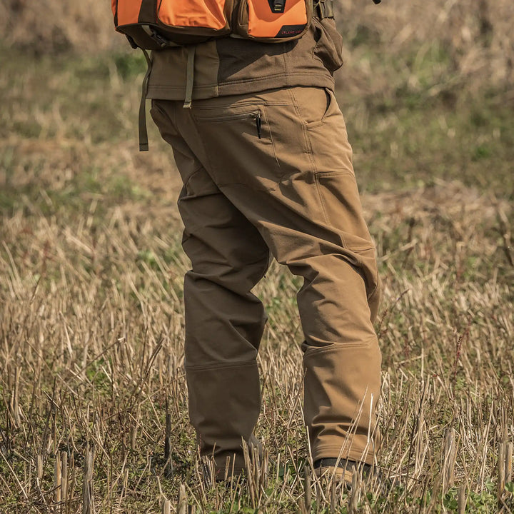 Person wearing tan Badlands pants and an orange backpack stands in a grassy field. Badlands skull logo visible on gear.