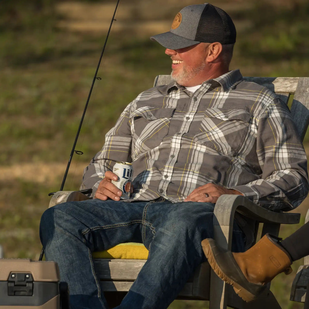 Man in plaid shirt and Badlands hat with skull logo relaxes outdoors, holding a can of beer beside fishing gear.