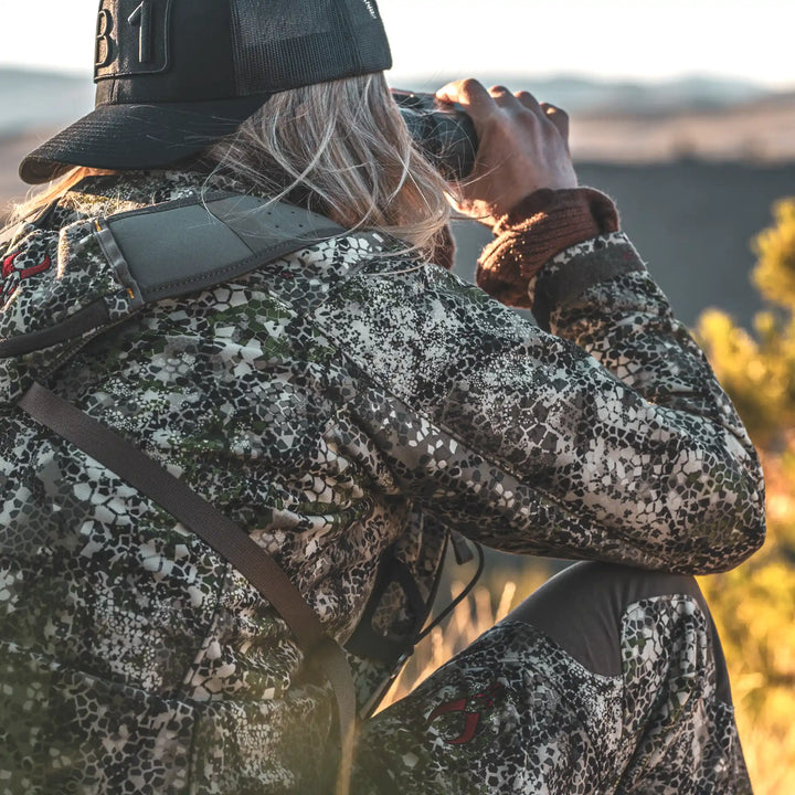 Person in Badlands camo gear with visible skull logo, using binoculars outdoors, seated in natural landscape.