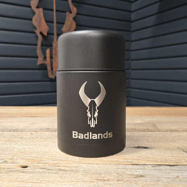 Black Badlands thermos with skull logo and text, placed on a wooden table with a dark paneled wall in the background.