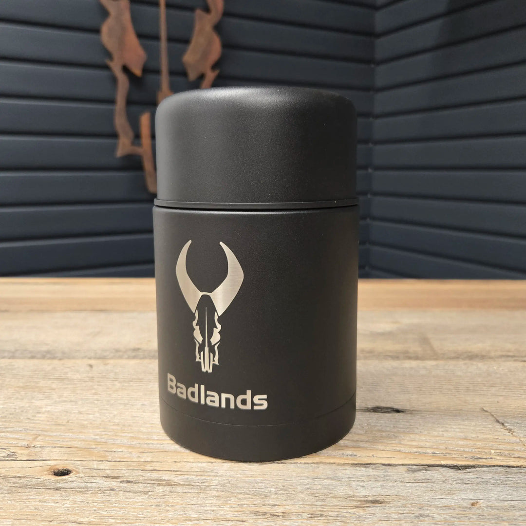 Black Badlands thermos with skull logo on a wooden table, dark paneled wall and metal logo in the background.