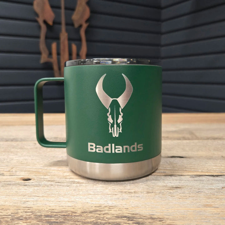 Green Badlands mug with silver skull logo and text, on a wooden table with dark panel wall in the background.